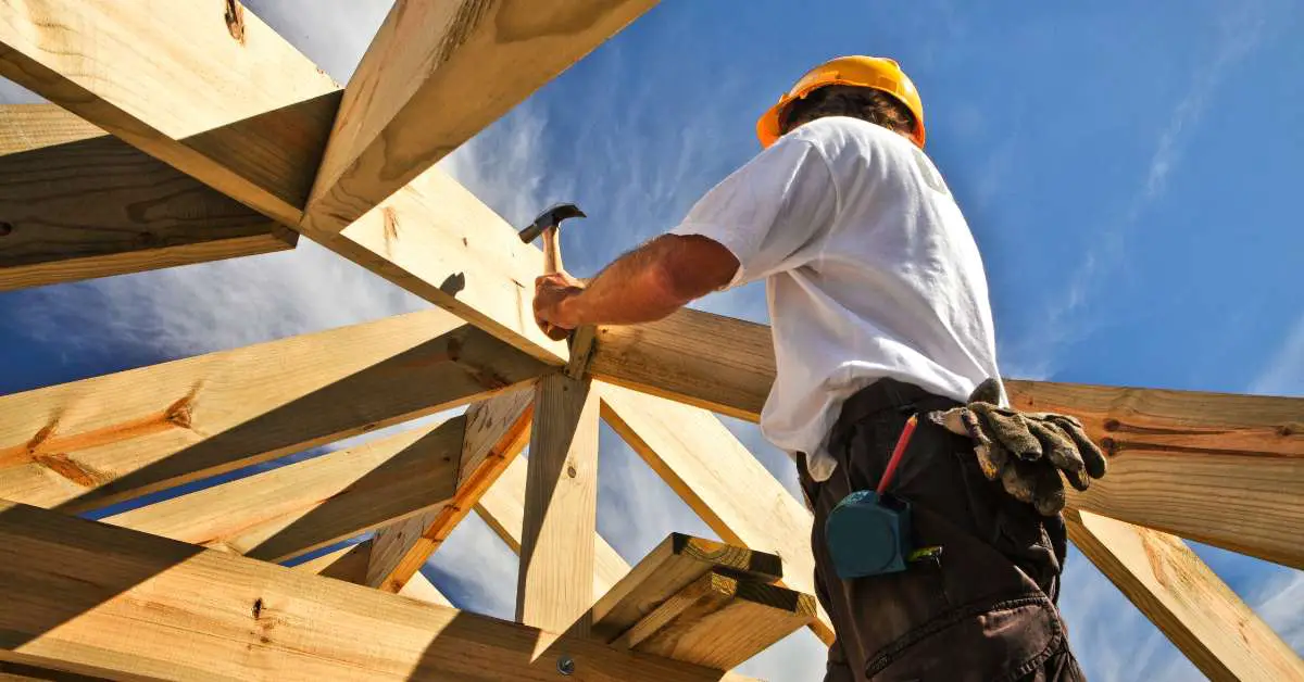 a worker poised to hammer a nail into a wooden plank, seemingly part of a house foundation