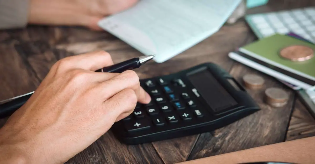 close-up of a hand holding a black pen, about to press a button on a calculator placed on a dark wood table, surrounded by coins, notebooks, and various stationery items