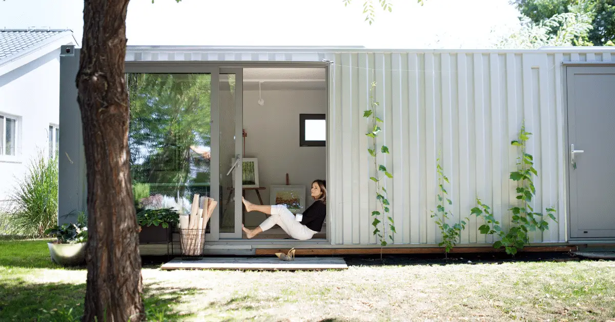 woman sitting on the floor of her insulated shipping container home, leaning against an open wide glass door, looking to the left