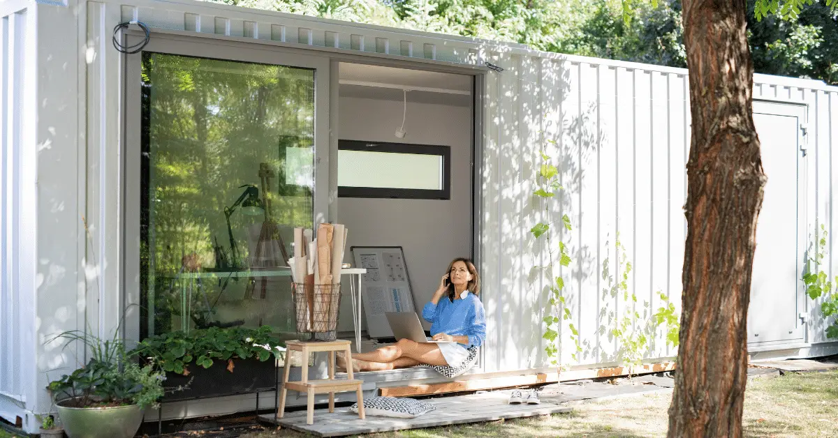 woman seated on the floor of her container home, engaged in a phone call, with an open MacBook on her lap and her spacious glass door ajar