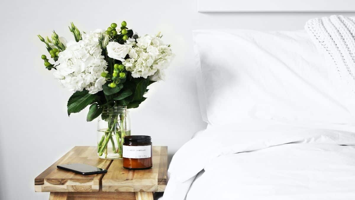 A vase of flower, a candle, and a phone on top of a table beside the bed in an apartment bedroom.