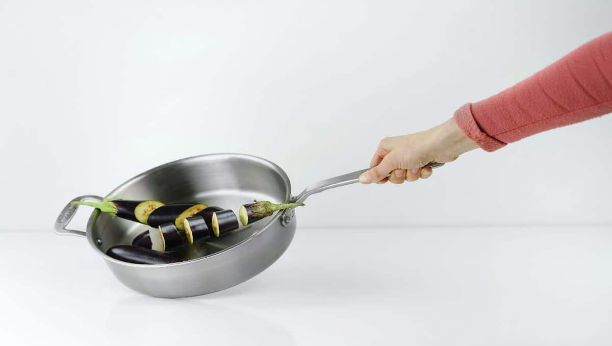 A hand holding a stainless steel pan with a sliced eggplant inside.